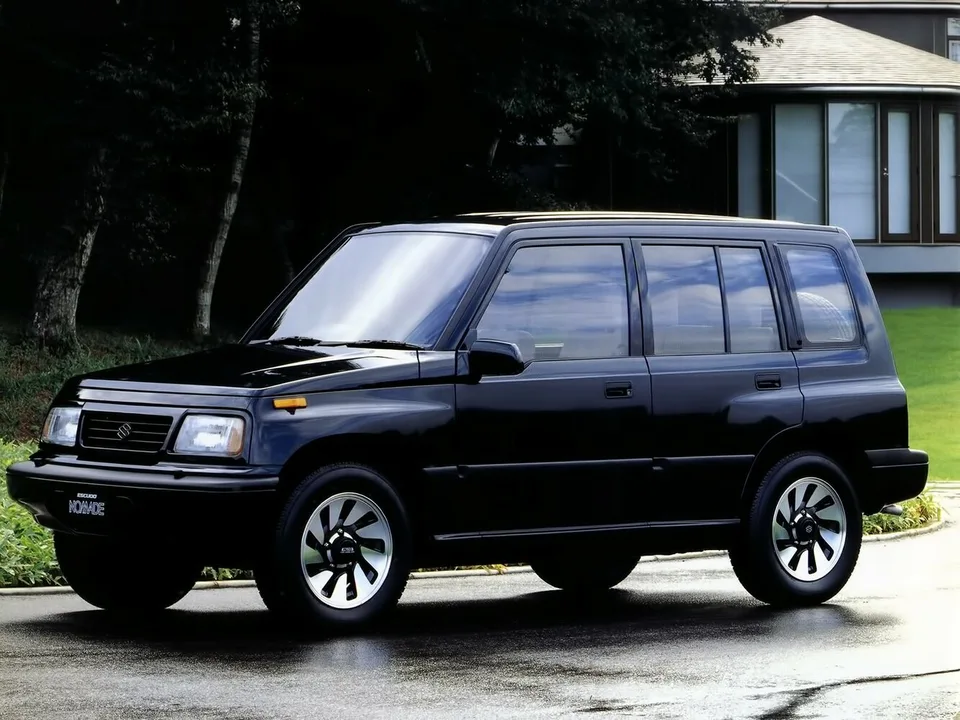 Suzuki Escudo Nomade первого поколения, чёрный пятидверный внедорожник на мокрой дороге у дома, вид спереди-сбоку слева