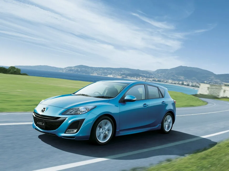 Mazda Axela второго поколения BL, голубой пятидверный хэтчбек в движении по приморской дороге, вид спереди-сбоку слева