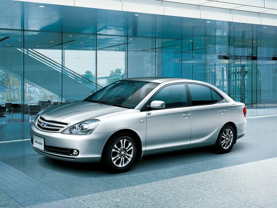 Toyota Allion первого поколения T240 после обновления, серебристый седан, вид спереди-сбоку слева, легкосплавные диски, фон — стеклянное здание
