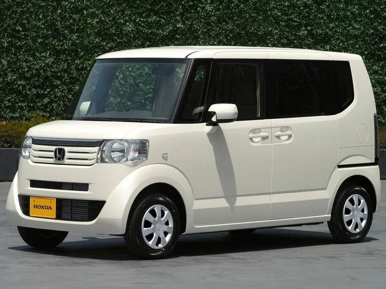 Honda N-BOX первого поколения, кремовый кузов, вид спереди-сбоку у живой изгороди, жёлтая табличка HONDA на бампере