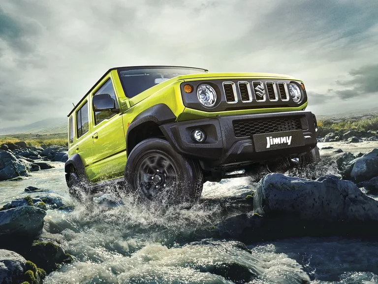 Пятидверный Suzuki Jimny с расширителями арок, зелёный кузов, едет по каменистому броду, хромированные планки решётки, табличка Jimny
