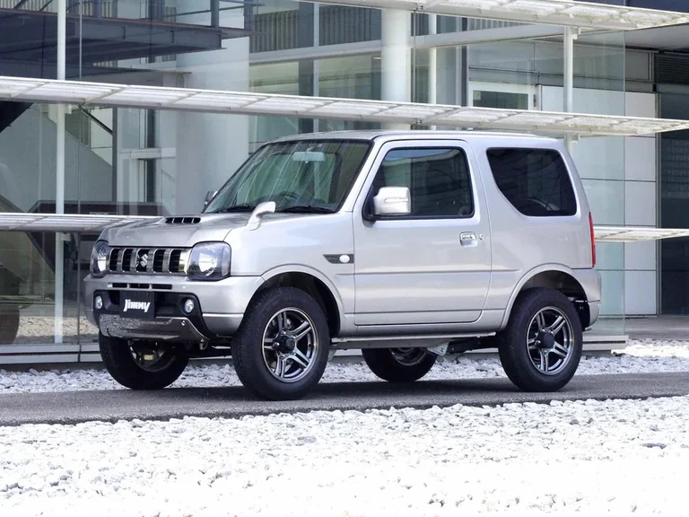 Suzuki Jimny JB23 позднего выпуска, серебристый трёхдверный кузов без расширителей арок на белом гравии у стеклянного здания, тёмная решётка с вертикальными прорезями