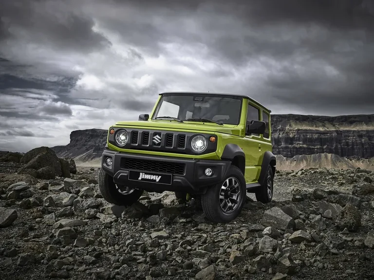 Suzuki Jimny четвёртого поколения с широким кузовом и чёрными расширителями арок, зелёный кузов, на каменистой площадке под грозовым небом, табличка Jimny