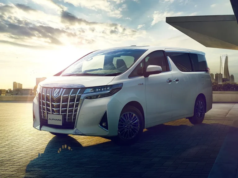 Toyota Alphard третьего поколения после обновления 2018 модельного года, белый минивэн на закате у современного здания, крупная решётка с вертикальными планками