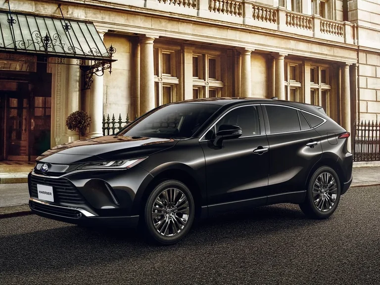 Toyota Harrier четвёртого поколения, чёрный кроссовер спереди-сбоку слева у здания с колоннами, табличка HARRIER на бампере, эмблема Toyota в синей окантовке